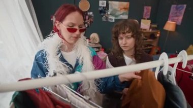 Kırmızı saçlı, beyaz saçlı, güneş gözlüklü, elbise askılarında koşuşturan bir kız. Evde bir bayan arkadaşıyla kıyafet seçerken.