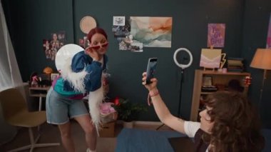 Kızıl saçlı, güneş gözlüğü takan, beyaz boa giyen, eşofman altı giyen ve evde akıllı telefon videosu kaydeden bayan arkadaşına poz veren bir kızın görüntüsünü büyüt.