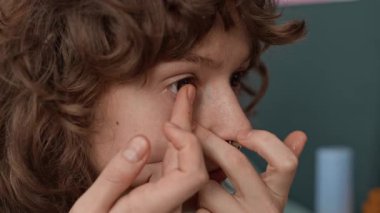Burnunda piercing olan genç bir kadının gözünün içine lens yerleştirirken ve aynada yüzünü incelerken yakın plan fotoğrafı.