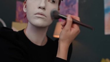 Yakından bakınca, kızıl saçlı, siyah kazaklı, beyaz boyalı, makyajlı, yüzlerine, kulaklarına ve boynuna sürdükten sonra siyah kazak giyen kız görülüyor.