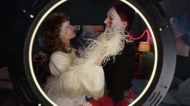 Yüzünde tüyler olan beyaz elbiseli kıvırcık saçlı bir kız ve pandomimci kız arkadaşı, şapka ve eldiven takmış ve halka ışığıyla video bloğu vuruyor.