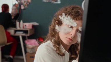 Dekoratif tüyleri yapıştırıp aynada kendine bakan ve kameraya poz veren odaklanmış kıvırcık saçlı kız.