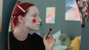 Evinde aynanın önünde palyaço makyajı yaparken beyaz taban üzerinde kırmızı daireler çizen konsantre genç bir kadının göğüsteki görüntüsü.