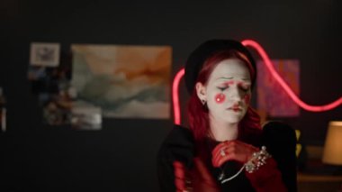 Siyah şapkalı, kırmızı eldivenli ve aksesuarlı, neon ışıklı odada kameraya poz veren çarpıcı palyaço makyajı yapan beyaz kadın portresi.