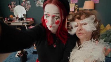 Kırmızı saçlı, pandomimci makyajlı ve şapkalı bir kız ve evinde dekoratif tüyler ve yapay elmaslar takan bir bayan arkadaşla kameraya poz veriyor.