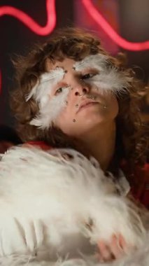 Beyaz tişörtlü, yaratıcı makyajlı, dekoratif tüylü, kameraya poz veren, kimliği belirsiz kırmızı eldivenli, elleri omuzlarını tutan bir kızın dikey portresi.