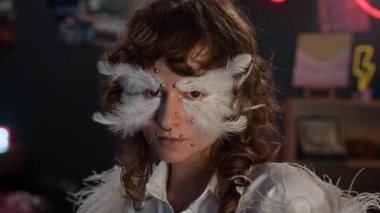 Kontak lensli ve septum piercingli UGC saçlı kızın beyaz gömlek, boa ve melek makyajı ve dekoratif tüyleri ile odasına inci çıkartmalar takıp kameraya poz verişi.