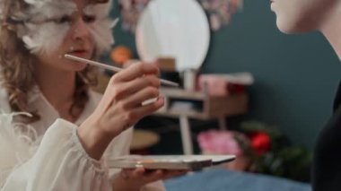 Z jenerasyonunun yakın plan çekimlerinde kız, evinde bayan arkadaşına palyaço makyajı yaparken elinde göz farı paleti ve fırça tutarak meleği oynuyor.