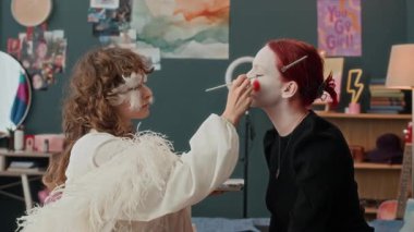 Yüzünde dekoratif tüyler olan, tiyatral beyaz bir kıyafet giymiş, kadın arkadaşına palyaço makyajı yapan kıvırcık saçlı genç bir kadın.