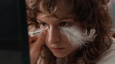 Makyaj yaparken yüzüne yapıştırdıktan sonra dekoratif tüy kesen burun ve kontak lensli kıvırcık saçlı kızın aynasına yakın çekim yap.