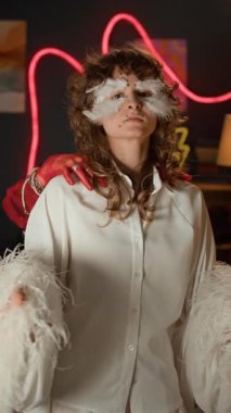 Kırmızı saçlı, palyaço makyajlı bir kızın dikey portresi beyaz elbiseli, yüzünde tüyler olan genç bir kadının arkasından beliriyor ve neon ışıklı odada birlikte poz veriyorlar.