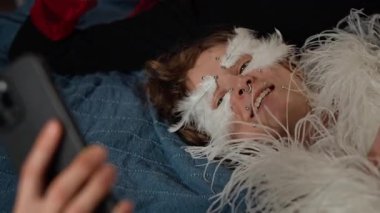 Evde kostüm partisi sırasında yatakta yatarken yaratıcı makyaj malzemesi giymiş ve akıllı telefon videosu çeken iki neşeli bayan arkadaşını yukarı kaldır.