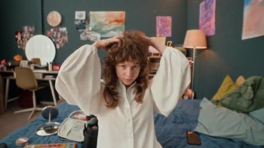 Beyaz bluz giymiş, saçlarını kontrol eden, şık yatak odasında kameraya bakan beyaz bir kadın.