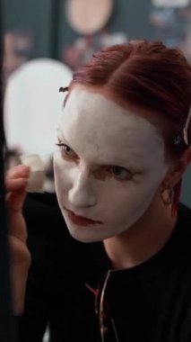 Kızıl saçlı, makyaj süngeri kullanan beyaz tabanlı genç bir kadın cadılar bayramı partisi ararken dikey görünümü