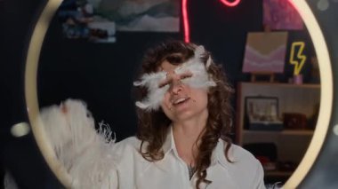 Pozitif kıvırcık saçlı, melek kıyafeti giyen, yüzünde inci çıkartmalar ve dekoratif tüyler olan bir kızın kamera karşısında kırmızı lambayla video bloğunda konuştuğu pozitifti.