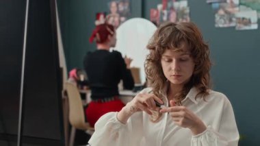 Göğüs hizasında beyaz bluzlu kız kozmetik yapıştırıcı açıyor ve dekoratif tüy alıyor. Arkadaşı arka plandaki makyaj masasında makyaj yapıyor.