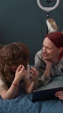 Dikey olarak iki kaygısız genç kadının yatakta birlikte yatması ve eğlenmesi, kızıl saçlı kızın dijital tablet tutması.