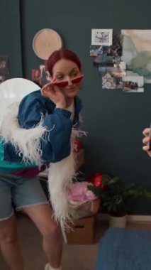 Evinde beyaz boa ile poz veren, kızıl saçlı, neşeli bir kadının akıllı telefon videosunu çeken, tanınmayan bir kızın elleri dikey.