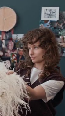 Beyaz t-shirt ve kahverengi yünlü yelek giyen beyaz kıvırcık saçlı genç bir kadının, evden kıyafet seçerken bilinmeyen bir arkadaştan pembe aksesuar alışının dikey görüntüsü.