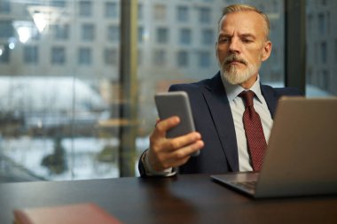 Ofiste dizüstü bilgisayarla çalışırken beyaz takım elbiseli bir iş adamının akıllı telefon ekranına baktığı orta boy bir fotoğraf.