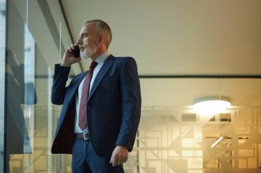 Olgun Kafkas işadamı modern ofiste duruyor, panoramik pencereden dışarı bakıyor ve telefonla konuşuyor, boşluğu kopyalıyor.