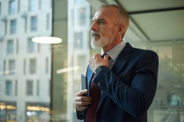 Orta yaşlı beyaz iş adamının resmi takım elbise giyip, kravatını düzeltirken, ofiste dikilirken orta yaşlı bir fotoğrafı.