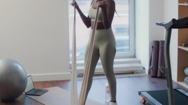 Afro-Amerikalı bir kadının paspasın üzerinde durup vücut egzersizi sırasında direnç bandıyla biseps kıvrımlarını çekerken.