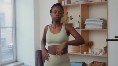 Genç Afrikalı Amerikalı kadın minimalist ev spor salonunda antrenman yapmadan önce gövde dönüşleri ve yan kıvrımlar yapıyor.