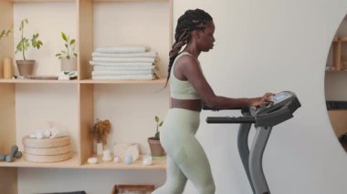 Spor giyimli genç siyahi kadının koşu bandı ayarlarını seçerken ve minimalist ev jimnastik salonunda koşmaya başlarken yan görüntüsü