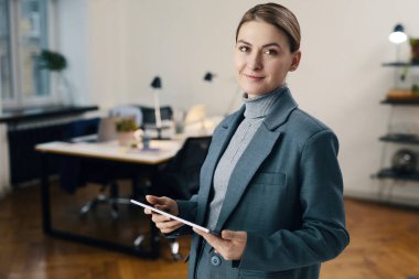 Ofiste dikilip dijital tablet üzerinde çalışırken kameraya bakan takım elbiseli genç iş kadınının portresi