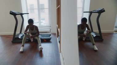 Spor salonundaki koşu bandında oturan Afrikalı Amerikalı kadın, terini alnından siliyor ve kardiyo antrenmanını yaparken şişeden su içiyor.