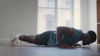 Aktif giyimli atletik siyah bir adam, modern ev spor salonunda, geniş pencereli spor salonunda egzersiz minderi üzerinde şınav çekiyor.