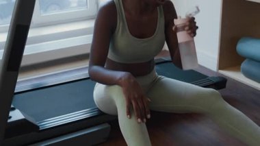 Koşu bandında oturan, şişeden su içen ve spor salonundaki antrenmandan sonra alnından ter silen yorgun siyahi kadını yukarı kaldır.