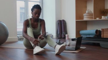 Spor kıyafetli genç bir kadın spor minderinde dizüstü bilgisayarla oturuyor, ayakkabı bağcıklarını bağlıyor ve evde antrenmana hazırlanıyor.