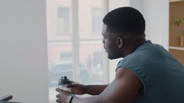 Koşu bandına yaslanmış yorgun siyah bir adam, su içiyor ve evde spor yaptıktan sonra terini alnından siliyor.