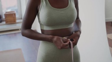Aktif giyimli genç siyahi bir kadının aynanın önünde durup, belini bantla ölçüp, evde antrenman yaptıktan sonra yansımasına bakarken görüntüsünü kaldır.