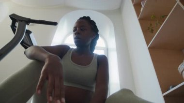 Yorgun siyah bir kadının evde oturup şişeden su içmesi ve koşu bandı egzersizinden sonra nefes almaya çalışması.