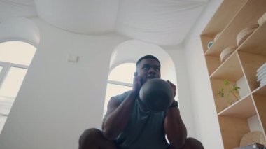 Spor elbiseli, kettlebell 'i iki koluyla tutan ve spor salonunda ağırlık çalışırken çömelme hareketleri yapan atletik siyahi bir adamın düşük açılı görüntüsü.