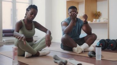 Spor giyimli genç bir çift spor salonunda bacak bacak üstüne atarak spor minderleri üzerinde oturur, su içer ve sohbet ederler.