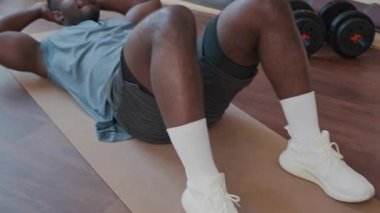 Aktif giyimli genç siyah bir adam egzersiz minderinde mekik çekerken evde spor yaparken karın kaslarını güçlendiriyor.