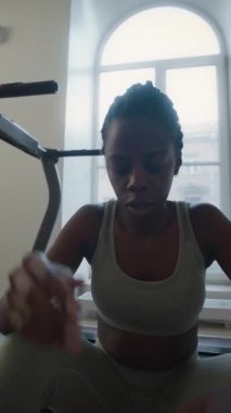 Yorgun Afrika kökenli Amerikalı bir kadının koşu bandında oturduğu, nefesini tuttuğu ve yüzündeki teri havluyla silerken yoğun ev egzersizinden sonra dinlendiği dikey bir fotoğraf.