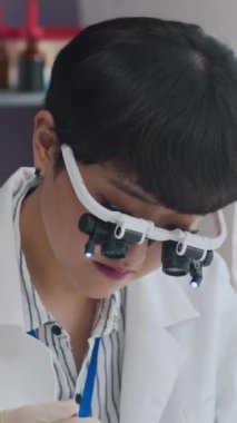 Laboratuvar önlüğü giymiş, büyüteç takmış, petri kabına pembe reaktör ekleyen Asyalı bilim adamının dikey görüntüsü.