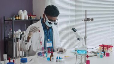 Laboratuvar önlüğü giyen, lateks eldiven takan ve petri kabı deneyi yaparken büyüteç takan Hintli bilim adamının statik çekimi.