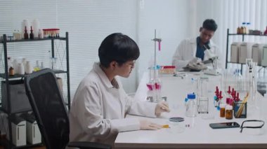 Hintli iş arkadaşı gözlem yazarken genç Asyalı laboratuvar çalışanı turnusol testi ile kimyasal reaksiyonu kontrol ediyor.