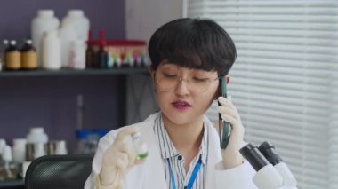 Asya 'lı kadın bilim adamı patronuyla akıllı telefonda konuşurken elinde aşı olan küçük bir şişeyi kıpırdatırken orta boy bir fotoğraf.
