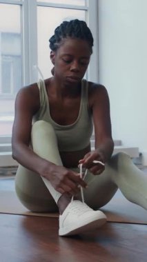 Genç siyahi bir kadının spor salonunda egzersiz minderinde otururken, antrenmandan önce ayakkabılarını bağlarken dikey pozu.