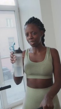 Genç Afrikalı Amerikalı bir kadının elinde bir şişe su tutarken omuz hizasında, spor salonundaki antremandan sonra partneriyle gülümseyip sohbet ederken.