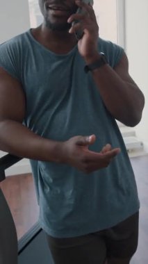 Spor salonunda koşu bandına yaslanan ve spor yaptıktan sonra cep telefonuyla sohbet eden neşeli siyahi adamın dikey çekimi.