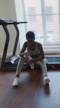 Spor giyim kuşağında yorgun siyah bir kadının spor salonunda koşu bandında oturması, yoğun idmandan sonra terini yüzünden silmesi ve su içmesi.