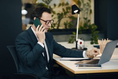 İşadamı iş yerindeki bilgisayarında çalışırken telefonda konuşuyor.